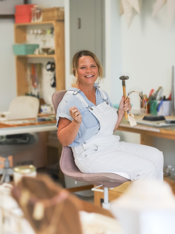 Female jewelry designer at work bench wearing white overalls and a blue shirt holding some of her tools.