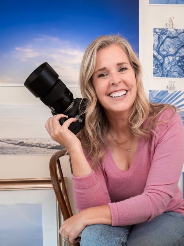 Paula McIntosh, fine art photographer and owner of PJM Photography, smiling while holding a professional camera in front of a wall displaying her ocean and landscape prints.
