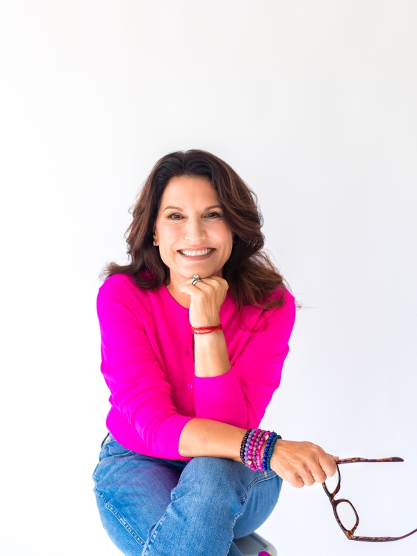 Julie Kaminski, board-certified health coach in Charleston, SC, smiling in a bright pink top with bracelets, seated in a relaxed, approachable pose.