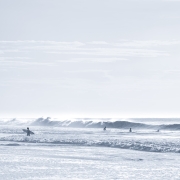 Fine art photograph by Paula McIntosh of PJM Photography capturing surfers in the gentle morning light, wading and waiting among rolling waves under a soft, silvery-blue sky.