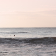 Fine art photograph by Paula McIntosh of PJM Photography showing a lone surfer floating on calm waves under a soft pink sky, evoking a quiet moment of stillness and reflection.