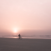 Fine art photograph by Paula McIntosh of PJM Photography showing a lone cyclist riding along the beach at sunrise under a soft pink sky and calm ocean horizon.