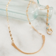 A gold hammered curved bar pendant on the Goldfield chain displayed on a blue green colored vintage bottle