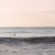 Fine art photograph by Paula McIntosh of PJM Photography showing a lone surfer floating on calm waves under a soft pink sky, evoking a quiet moment of stillness and reflection.