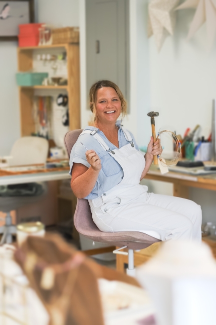 Female jewelry designer at work bench wearing white overalls and a blue shirt holding some of her tools.