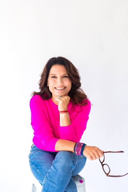 Julie Kaminski, board-certified health coach in Charleston, SC, smiling in a bright pink top with bracelets, seated in a relaxed, approachable pose.
